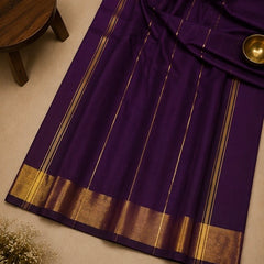 A deep purple saree with elegant gold stripes and a wide gold zari border, laid out on a neutral beige surface with a small wooden stool and a brass bowl as props.