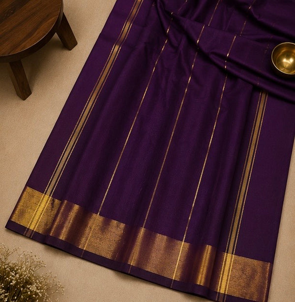 A deep purple saree with elegant gold stripes and a wide gold zari border, laid out on a neutral beige surface with a small wooden stool and a brass bowl as props.