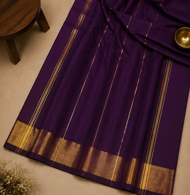 A deep purple saree with elegant gold stripes and a wide gold zari border, laid out on a neutral beige surface with a small wooden stool and a brass bowl as props.